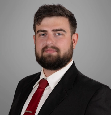 Christian Brooks, criminal defense attorney in Greensboro, NC, at Dummit Fradin law firm, wearing a black suit, white shirt, and red tie, posed against a gray background.